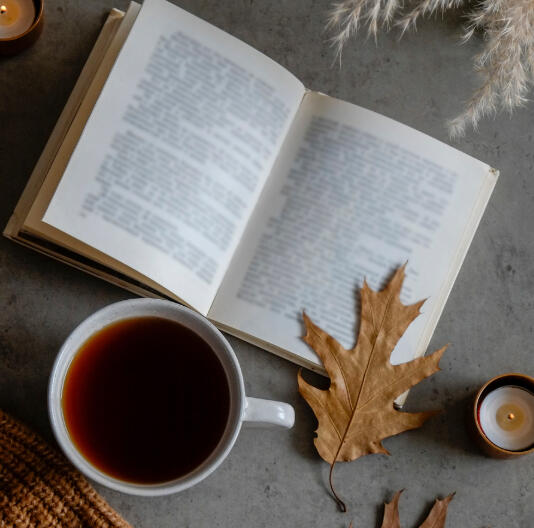 An open book with an orange oak leaf resting on the page, a candle off to the left side of the book, and a cup of tea