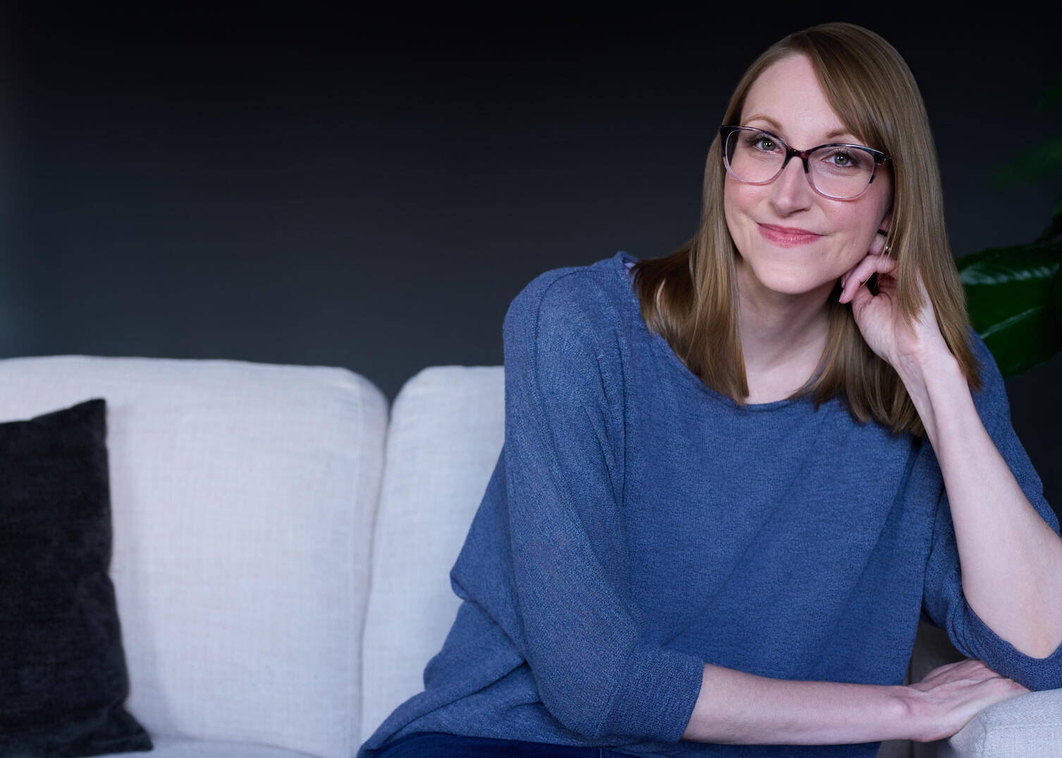 A woman with blonde hair and glasses sits on a white couch, leaning on her hand and smiling